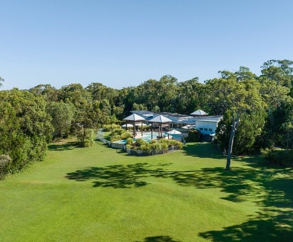 Aerial view of a spacious, modern home surrounded by lush greenery and a swimming pool, set against a clear blue sky.