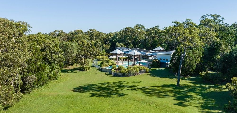 Aerial view of a spacious, modern home surrounded by lush greenery and a swimming pool, set against a clear blue sky.