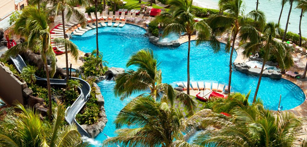 Aerial view of a luxurious resort pool surrounded by palm trees, featuring slides and sun loungers, with the ocean in the background.