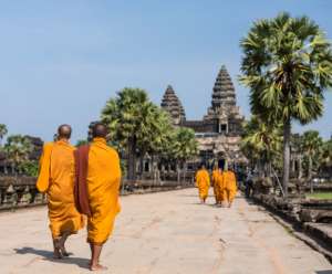 Monks in orange robes walk towards Angkor Wat, framed by palm trees and a clear blue sky.