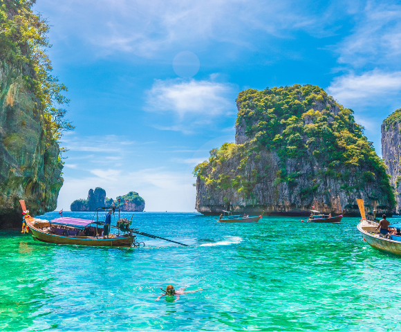 Four traditional wooden longtail boats on a serene beach with crystal-clear water, against a backdrop of a rocky island and clear blue sky.