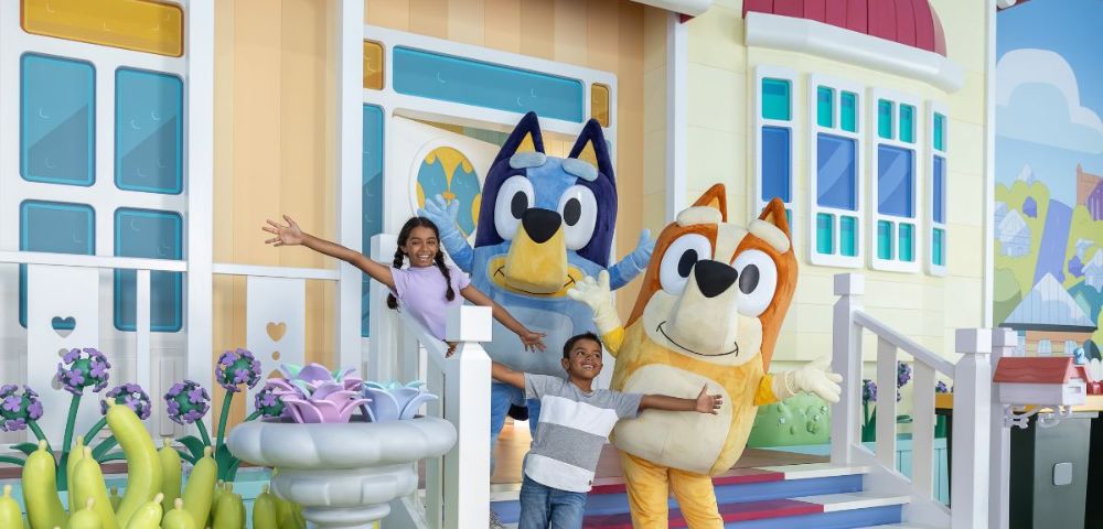 Two colorful costumed characters greeting children in front of a cheerful, cartoonish house backdrop filled with flowers and greenery.