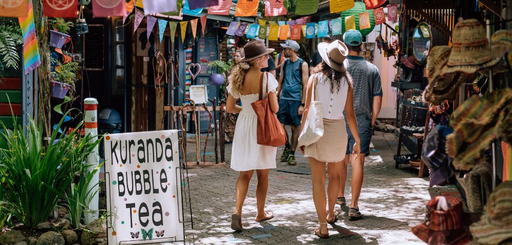 Three people stroll through a vibrant market, adorned with colorful flags and banners, beside a sign for 