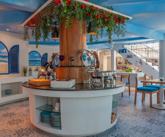 Stylish dining area with a circular buffet, blue decor, lush greenery, and beach-themed wall art, creating a vibrant and inviting atmosphere.