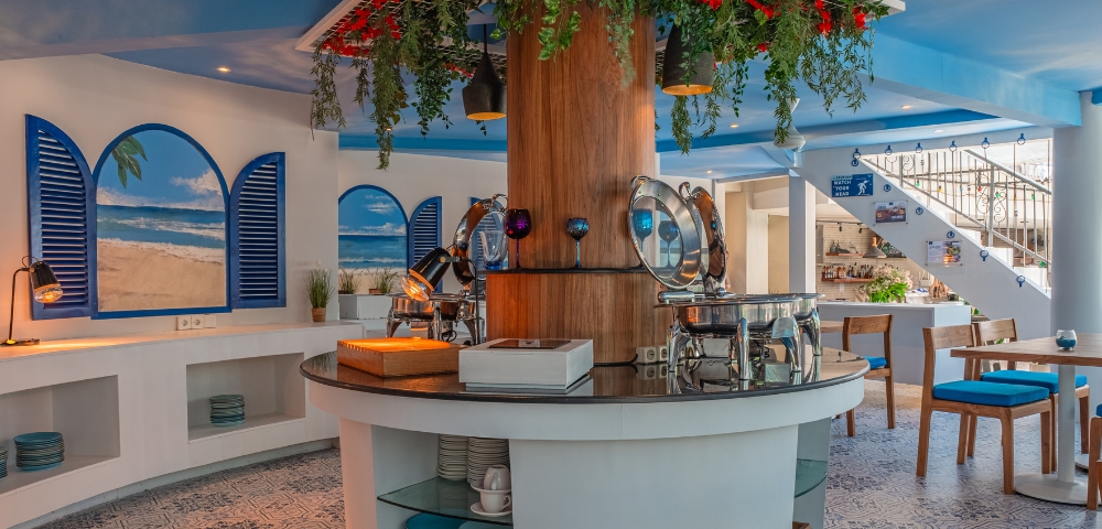 Stylish dining area with a circular buffet, blue decor, lush greenery, and beach-themed wall art, creating a vibrant and inviting atmosphere.