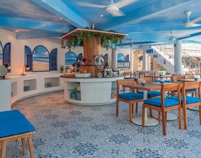 Bright and airy café interior with blue accents, wooden furniture, and a central bar, featuring decorative plants and artistic tile flooring.
