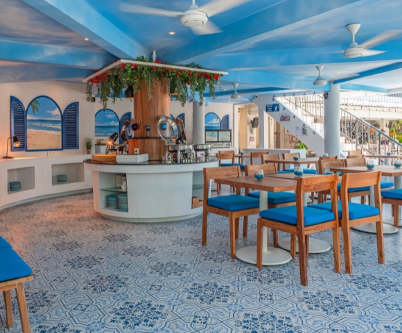 Bright and airy café interior with blue accents, wooden furniture, and a central bar, featuring decorative plants and artistic tile flooring.