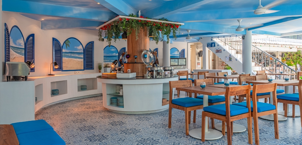 Bright and airy café interior with blue accents, wooden furniture, and a central bar, featuring decorative plants and artistic tile flooring.