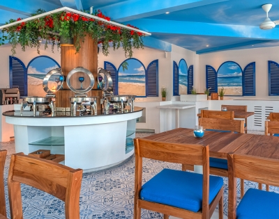 Bright and airy restaurant interior with blue accents, wooden tables, and a buffet counter surrounded by decorative plants.