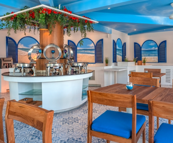 Bright and airy restaurant interior with blue accents, wooden tables, and a buffet counter surrounded by decorative plants.