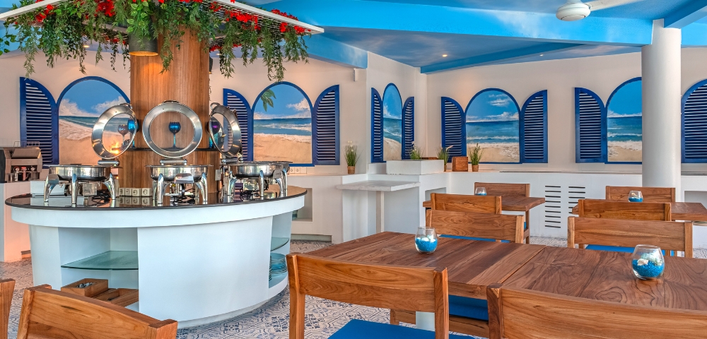 Bright and airy restaurant interior with blue accents, wooden tables, and a buffet counter surrounded by decorative plants.