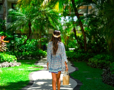 A woman in a floral dress walks along a winding path through a lush tropical garden, holding a woven basket.
