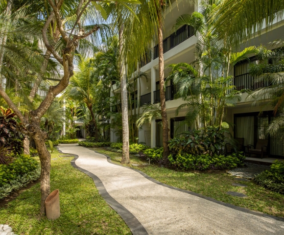 Lush tropical pathway winding between resort buildings, lined with palm trees and vibrant greenery under bright sunlight.