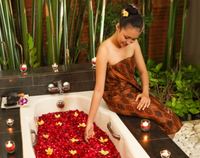 A woman sits in a luxurious bathtub filled with rose petals and floating flowers, surrounded by candles and lush greenery.