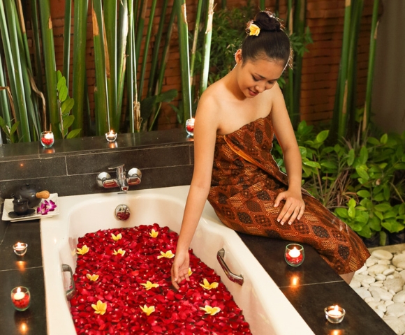 A woman sits in a luxurious bathtub filled with rose petals and floating flowers, surrounded by candles and lush greenery.