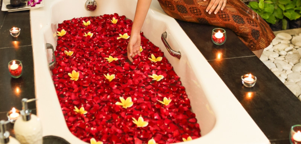 A woman sits in a luxurious bathtub filled with rose petals and floating flowers, surrounded by candles and lush greenery.