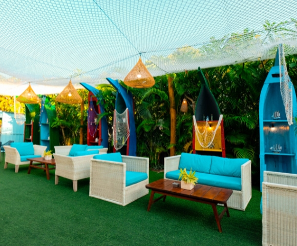 Cozy outdoor lounge with white wicker seating, vibrant blue cushions, surfboard decor, and greenery under a netted canopy.