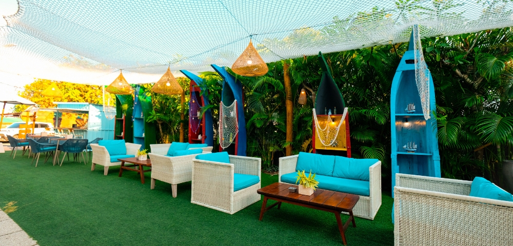 Cozy outdoor lounge with white wicker seating, vibrant blue cushions, surfboard decor, and greenery under a netted canopy.
