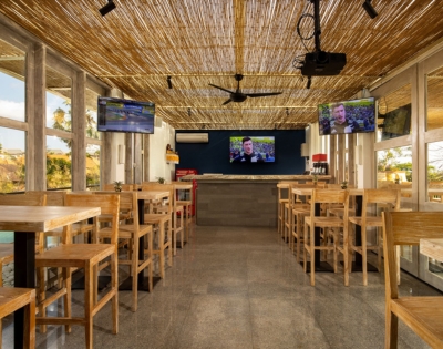 Bright, modern restaurant interior with wooden stools, a bar area, and multiple TVs displaying news, under a thatched roof.