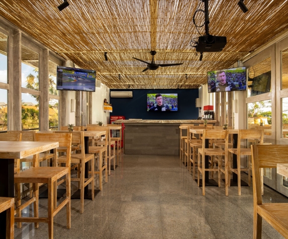 Bright, modern restaurant interior with wooden stools, a bar area, and multiple TVs displaying news, under a thatched roof.