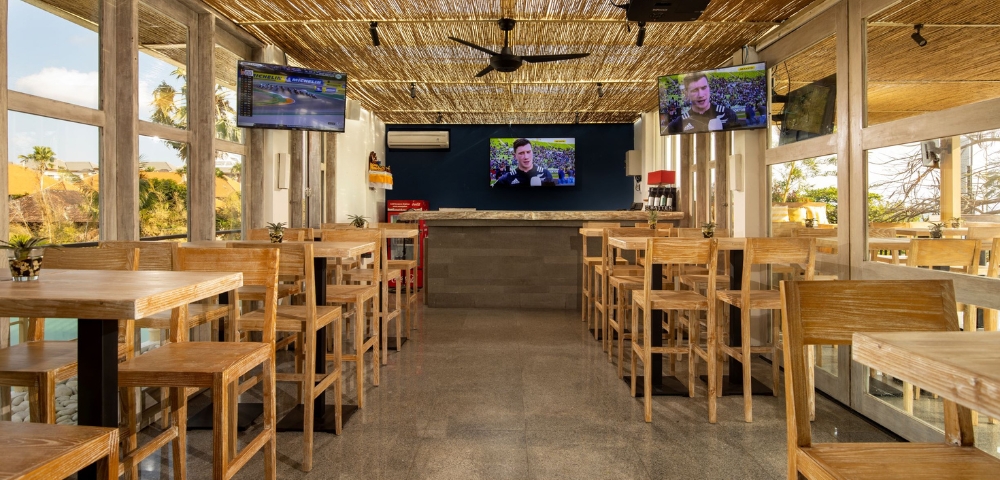 Bright, modern restaurant interior with wooden stools, a bar area, and multiple TVs displaying news, under a thatched roof.