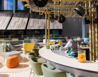 A bartender prepares drinks at a stylish rooftop bar featuring colorful seating and a modern design with natural light.