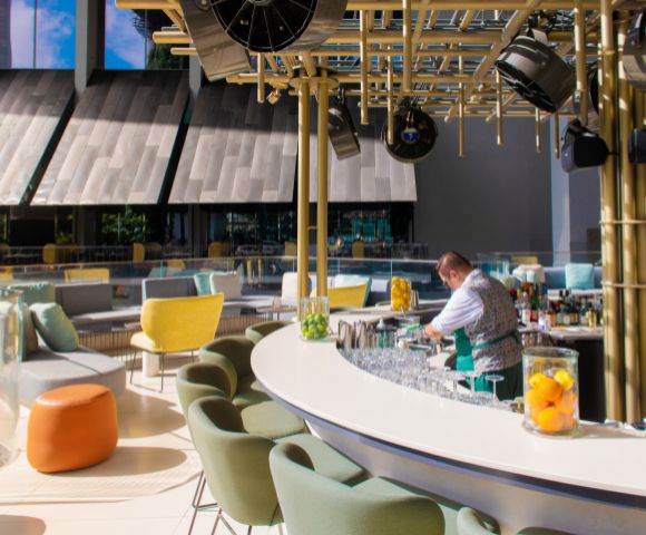 A bartender prepares drinks at a stylish rooftop bar featuring colorful seating and a modern design with natural light.