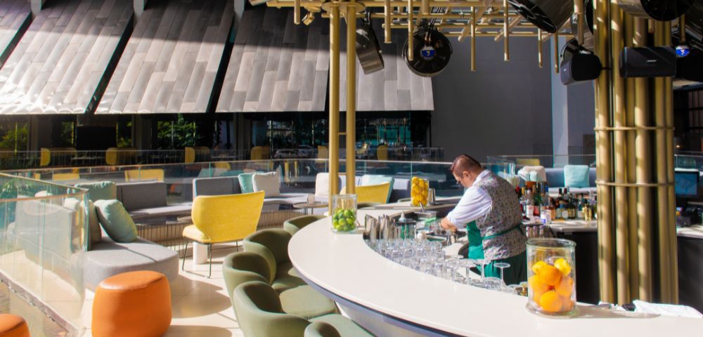 A bartender prepares drinks at a stylish rooftop bar featuring colorful seating and a modern design with natural light.