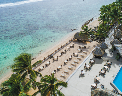 Aerial view of a tropical beach with palm trees, dining tables, thatched-roof huts, and a shimmering blue pool beside the ocean.