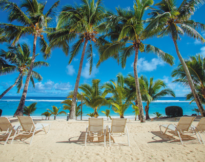 A serene beach scene with white lounge chairs under palm trees, facing turquoise waters and a clear blue sky.