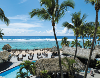 Tropical beach view featuring palm trees, a sparkling pool, and thatched-roof structures overlooking a serene ocean.