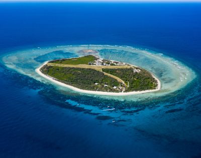 Aerial view of a small tropical island surrounded by clear blue waters and a coral reef, featuring lush greenery and a few buildings.