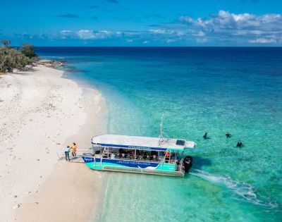 A boat anchored on a sandy shore, with people disembarking and swimmers enjoying the clear turquoise waters.