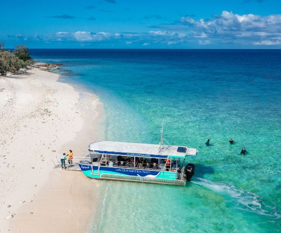 A boat anchored on a sandy shore, with people disembarking and swimmers enjoying the clear turquoise waters.