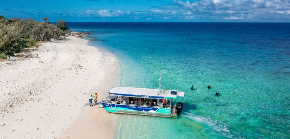 A boat anchored on a sandy shore, with people disembarking and swimmers enjoying the clear turquoise waters.