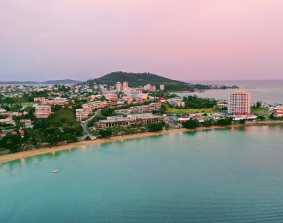 Aerial view of a coastal town at dusk, featuring sandy beaches, modern buildings, and lush hills along the shoreline.