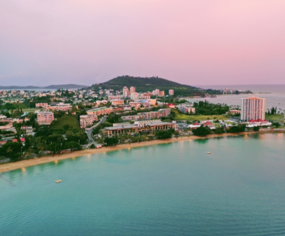 Aerial view of a coastal town at dusk, featuring sandy beaches, modern buildings, and lush hills along the shoreline.
