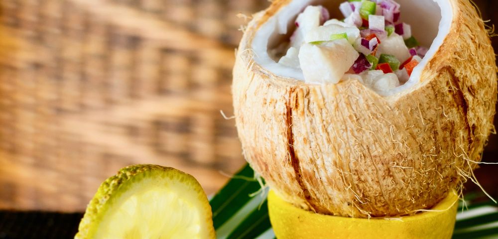 A coconut half filled with ceviche topped with chopped vegetables, served alongside lemon wedges and garnished with green leaves.