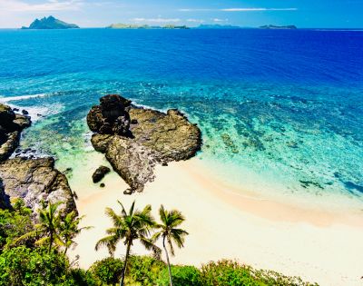 Idyllic beach with palm trees, rocky outcrops, and crystal-clear turquoise waters under a clear blue sky.