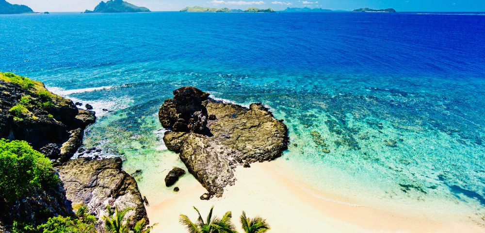 Idyllic beach with palm trees, rocky outcrops, and crystal-clear turquoise waters under a clear blue sky.