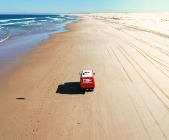 A red van drives along a deserted beach, with waves lapping at the shore and sand dunes in the background under a clear blue sky.