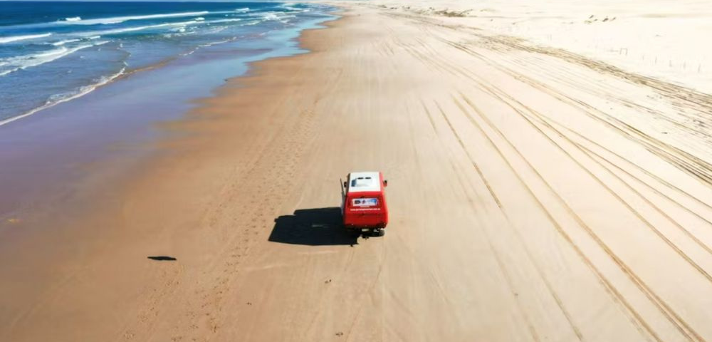 A red van drives along a deserted beach, with waves lapping at the shore and sand dunes in the background under a clear blue sky.