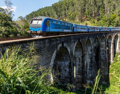 A blue train travels across a historic stone viaduct, surrounded by lush greenery and hills under a clear blue sky.