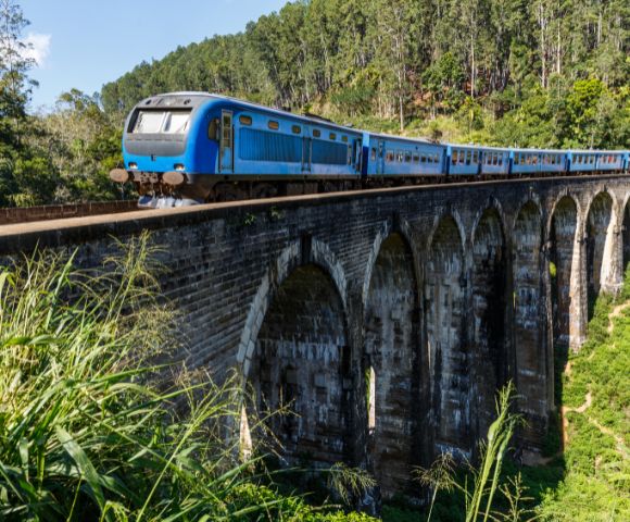 A blue train travels across a historic stone viaduct, surrounded by lush greenery and hills under a clear blue sky.
