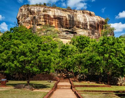 A stone formation rises above lush green trees under a bright blue sky, with a pathway leading to its base.