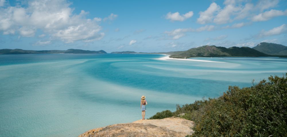 A woman stands on a rocky outcrop, gazing at a turquoise sea and sandy shores under a bright blue sky with scattered clouds.