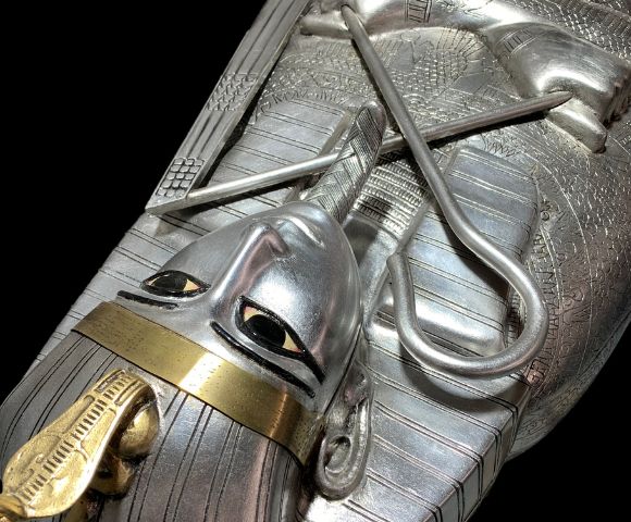 Close-up of a silver sarcophagus with a stylized human face, gold accents, and intricate engravings, showcasing ancient Egyptian artistry.