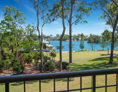 View from a balcony overlooking a serene lake, surrounded by lush greenery and trees, with a boat in the water under a clear blue sky.