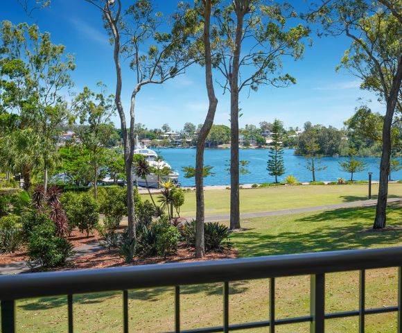 View from a balcony overlooking a serene lake, surrounded by lush greenery and trees, with a boat in the water under a clear blue sky.