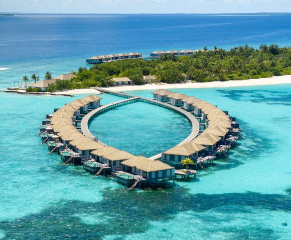 Aerial view of luxury overwater villas in a heart shape, surrounded by turquoise waters and lush tropical vegetation.